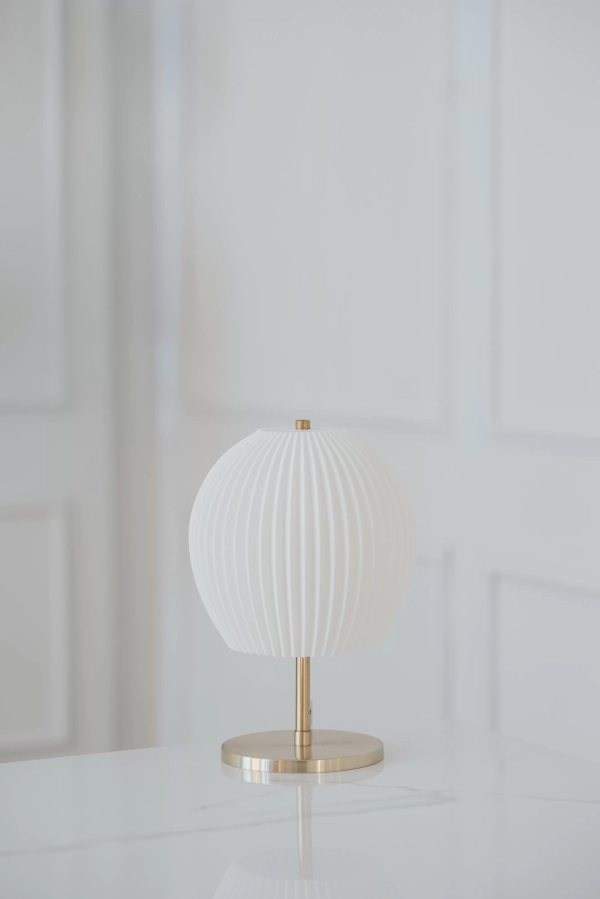 Lámpara de mesa Serena Mini. Fabricada con base metálica disponible en color bronce o negro, y pantalla en impresión 3D de color blanco. Estilo moderno.